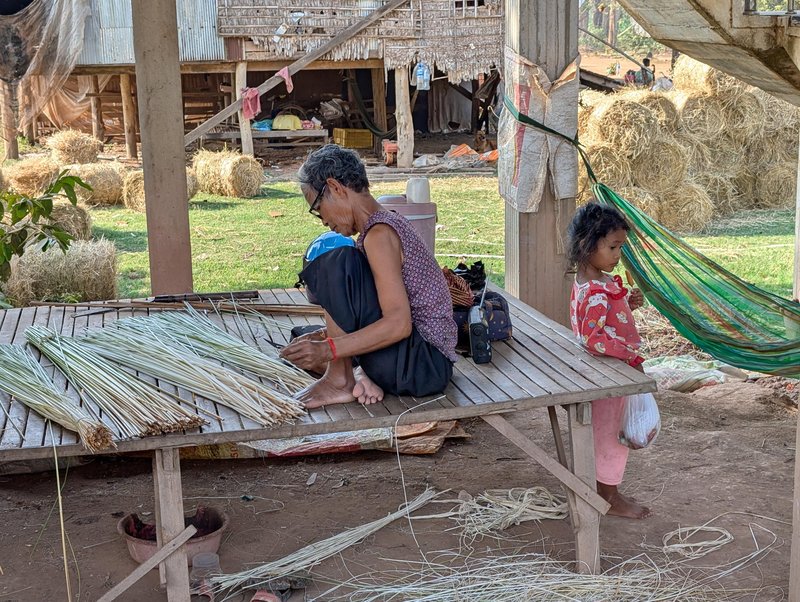 And older woman weaving some sort of grass with a small girl